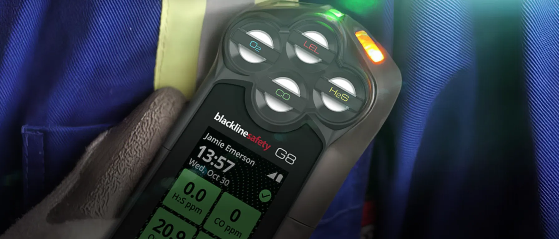 Handheld gas detector (Blackline safety G8) with four gas dials labeled O2, LEL, CO, and H2S, screen showing user name and gas readings in green panels, worn by a gloved hand against blue fabric background.